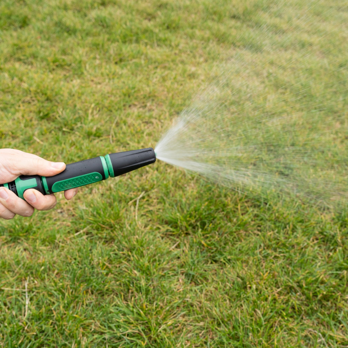 Hand hält die Gartenspritze am Schlauch und sprüht einen feinen Wasserfächer über den Rasen