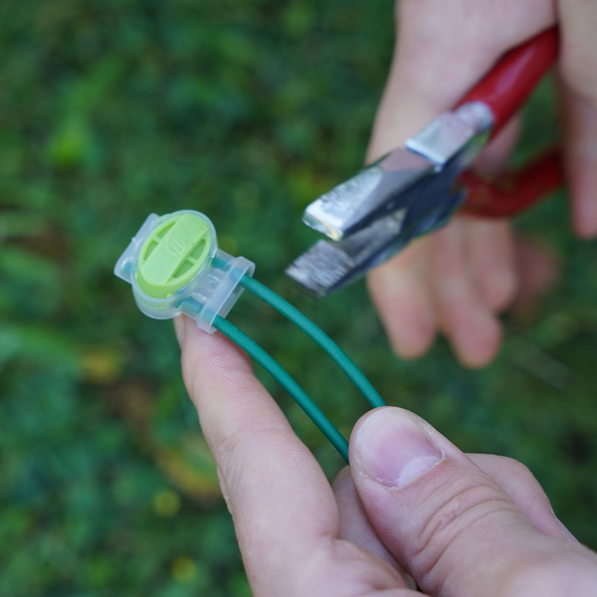 Zwei Kabelenden in einer grünen Verbindungsklemme, daneben eine Zange zum Zudrücken, im Garten